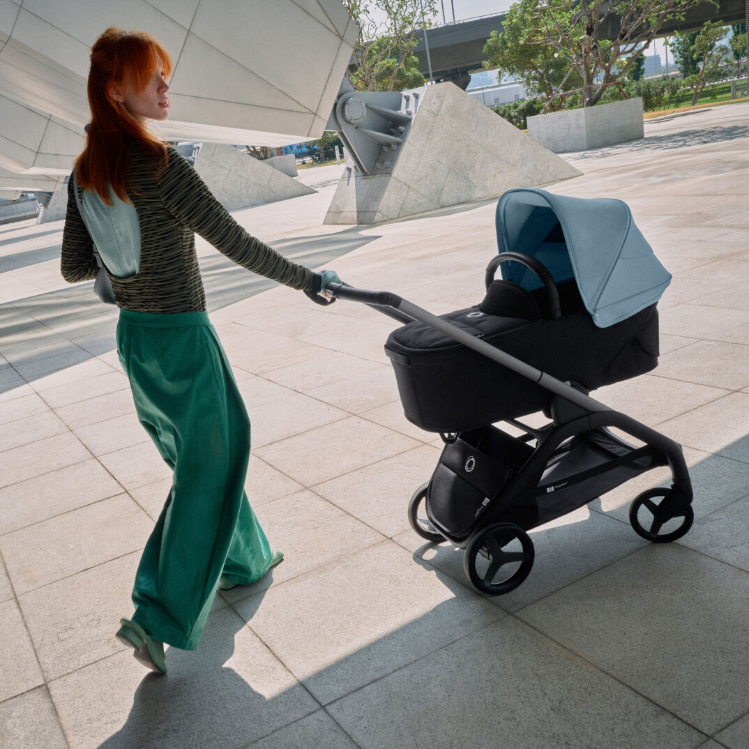 バガブー ドラゴンフライに赤ちゃんを乗せて、遠くに庭の見えるビルの前を散歩するスタイリッシュな母親。