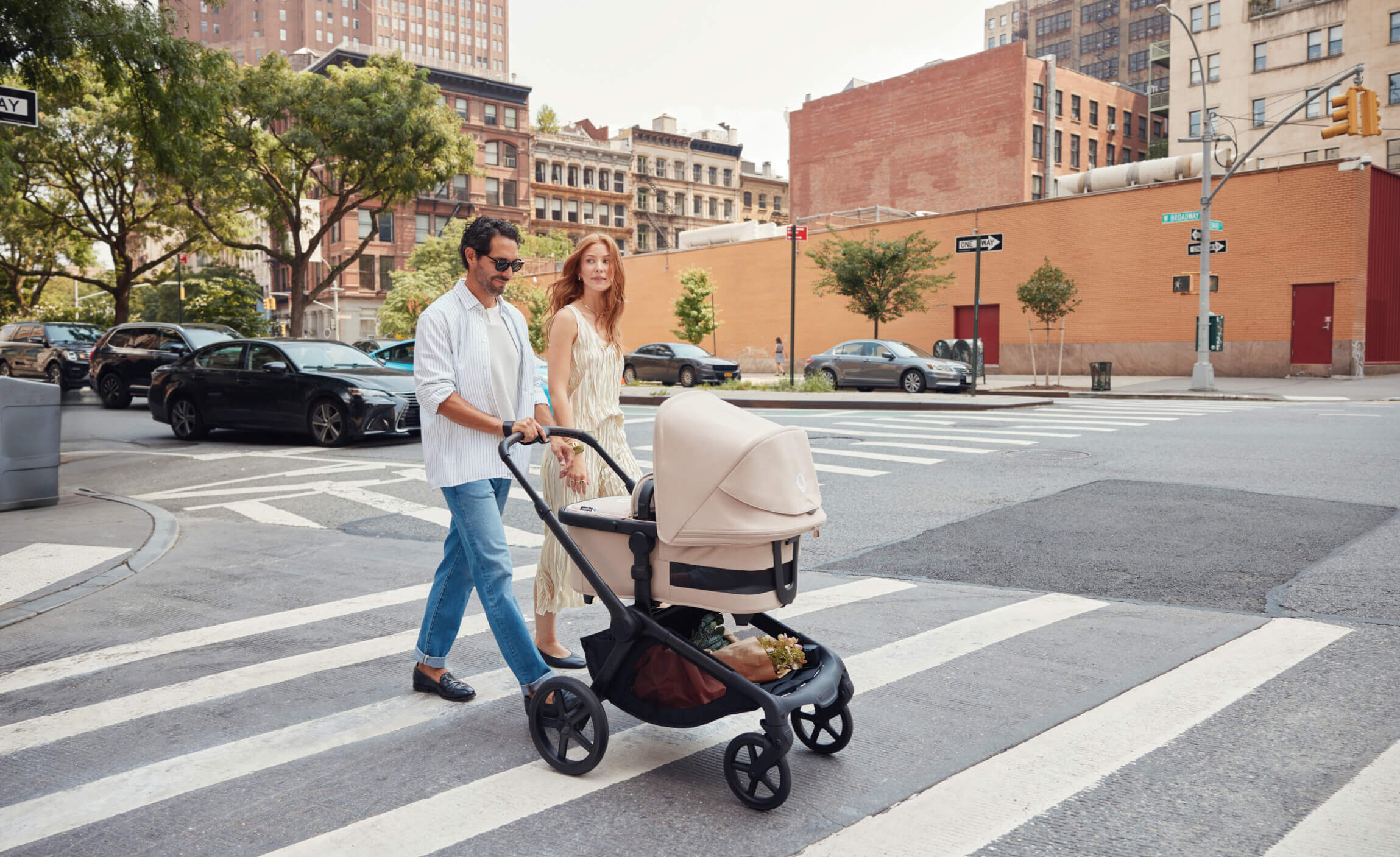 Two parents cross the street with their baby in a Bugaboo Kangaroo bassinet pram with Desert Taupe fabrics. They're in an urban setting with buildings and cars in the background.