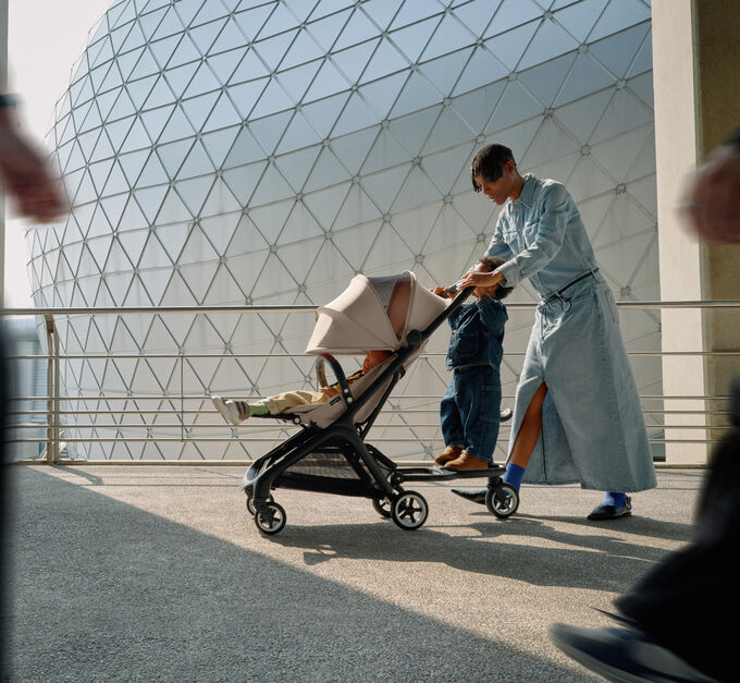 A stylish mom walks with her baby in a Bugaboo Butterfly travel stroller, while her toddler rides along on the wheeled board.