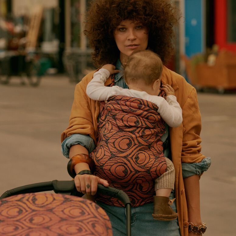 A stylish mom carries her baby in an Artipoppe Zeitgeist Baby Carrier. She’s holding onto a Bugaboo stroller with the Bugaboo X Artipoppe Limited Edition sun canopy. Behind them is a casual, urban setting.