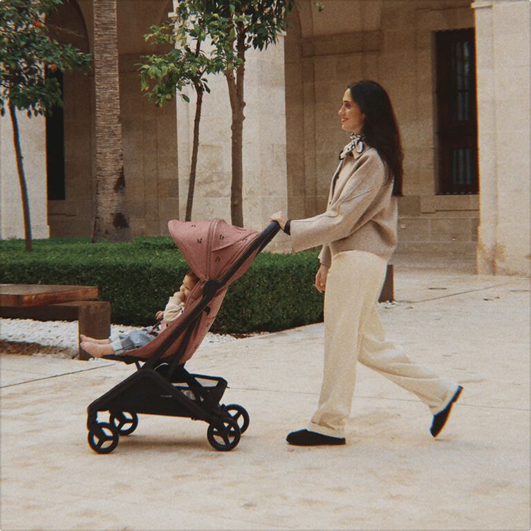A smiling mom walks with her baby sitting in a Bugaboo Butterfly 2 stroller. The stroller features limited-edition fabric embroidered with Konges Sløjd cherry motif. They’re in a green courtyard outside a stone-walled building.