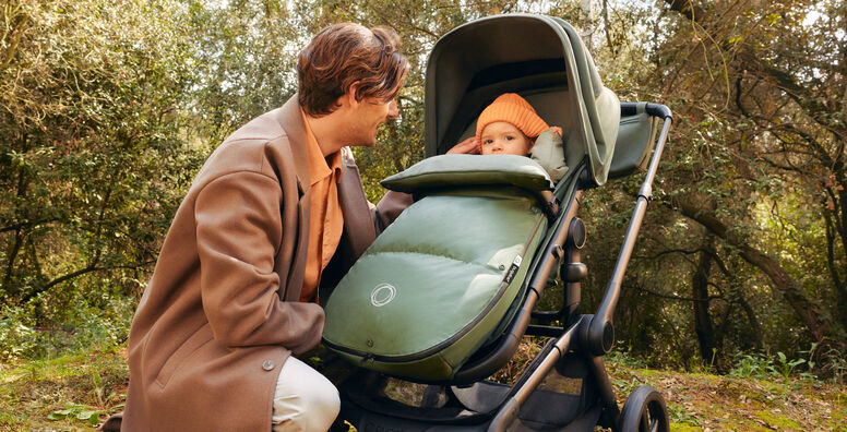 A parent pushes a Bugaboo Fox 5 Renew bassinet stroller with a Bugaboo wheeled board attached, while a child walks alongside.