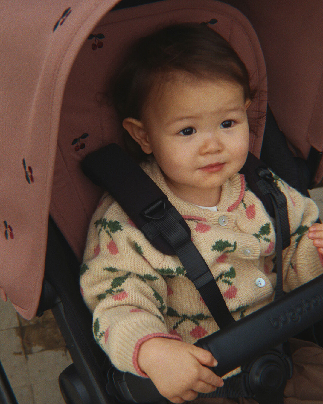 A close up of a Bugaboo x Konges Sløjd Limited Edition organizer, with cherry patterns embroidered on Dusty Pink fabric, worn over a beige coat.