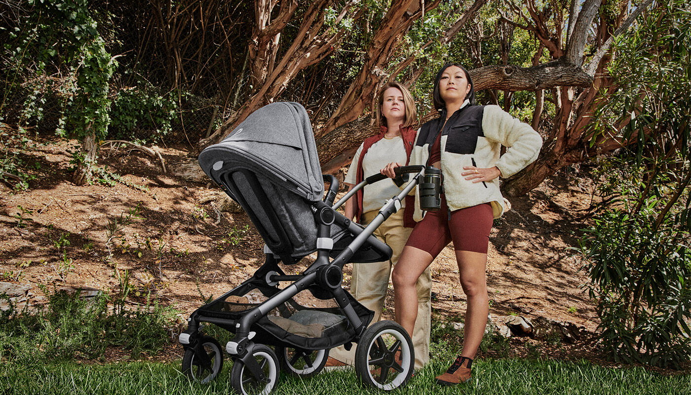 Two moms standing in a hero pose next to baby in a Bugaboo stroller in a nature park.