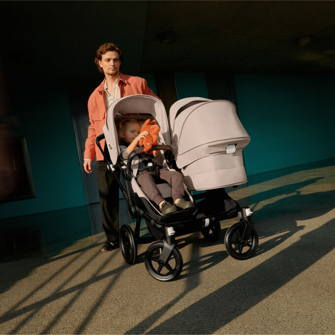 A dad makes a turn with a Bugaboo Donkey 5 Duo pram. His toddler is in the seat facing the world and holding a stuffed animal. The bassinet is facing the dad