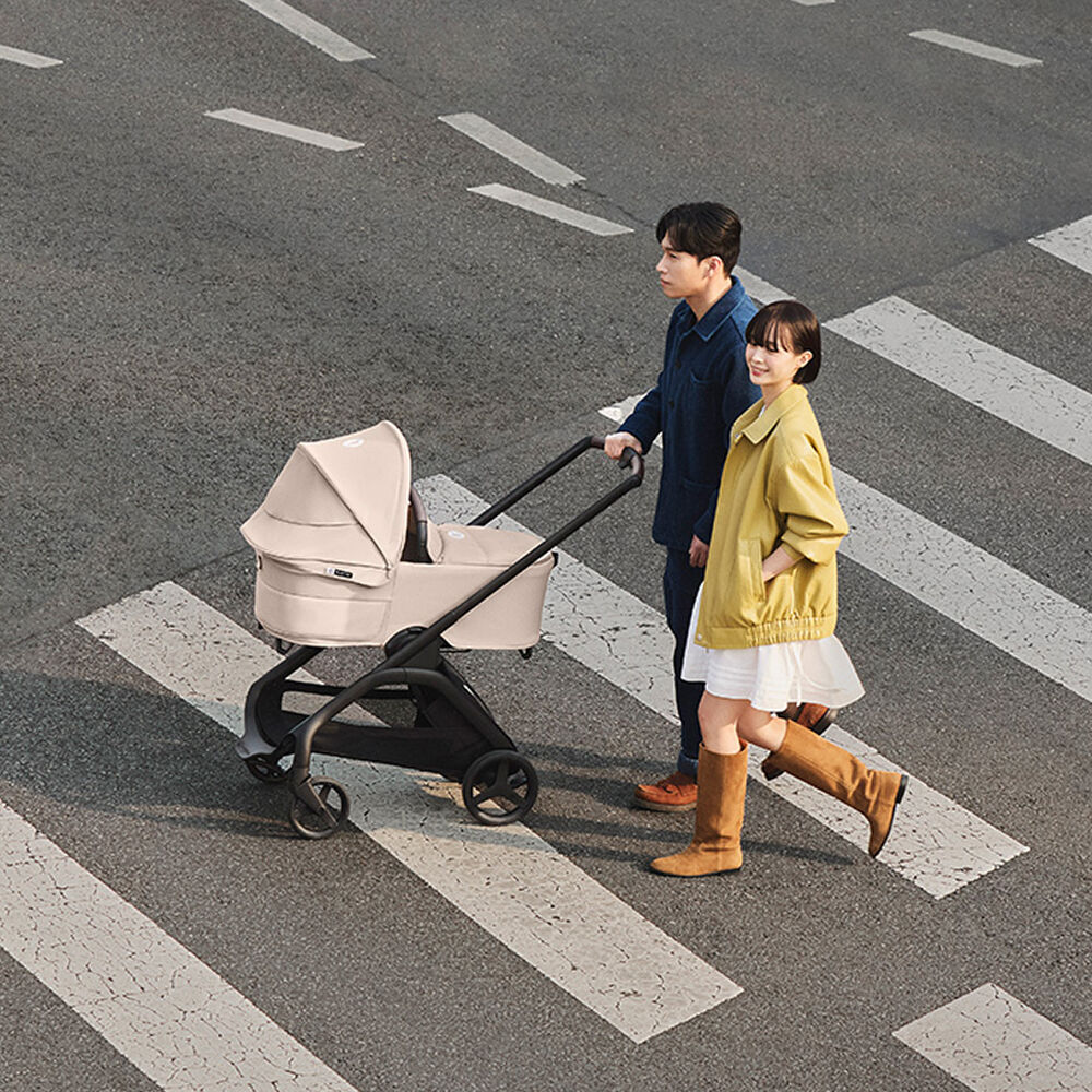 A couple walk with a Bugaboo Dragonfly Plus bassinet stroller across a wide striped crosswalk on an empty city street.