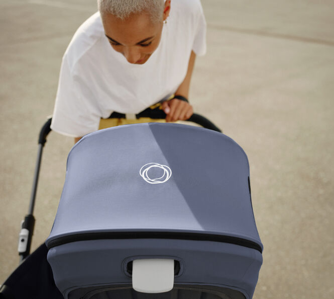 A mother leans over her Bugaboo pushchair to check on her baby. The Bugaboo pushchair has a Stormy Blue canopy, with a white Bugaboo logo in the centre.