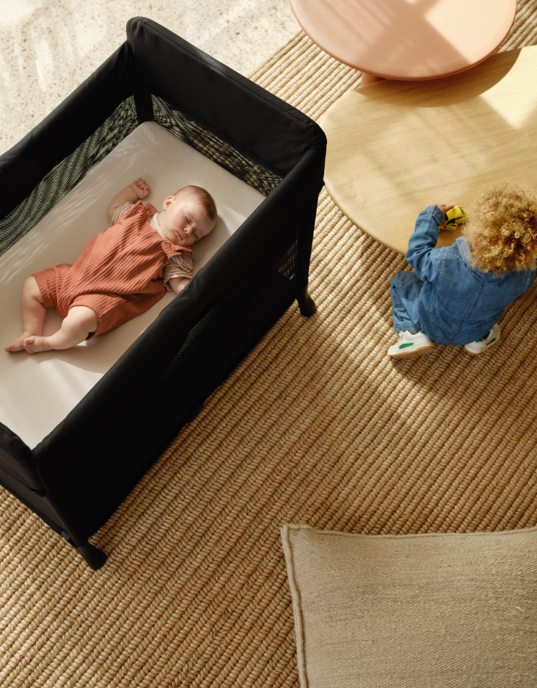 Top view of a baby napping peacefully inside a Bugaboo Stardust travel cot. Next to the travel cot, a toddler is playing at a low table.