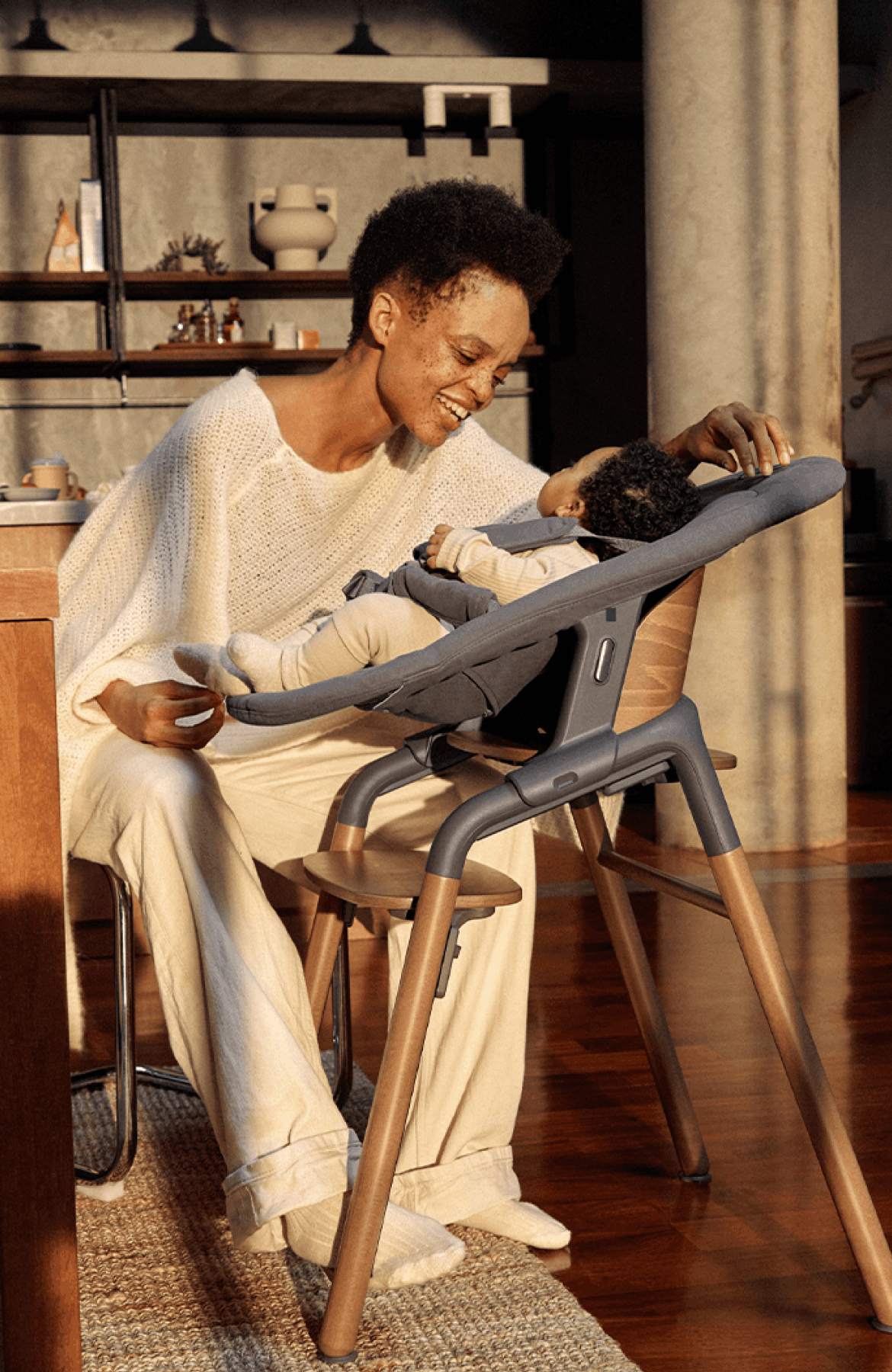 バガブージラフ ハイチェアに座る彼女の赤ちゃんと遊ぶ、カジュアルな服装の母親。 彼らは、暖かみのある照明に包まれたモダンなキッチンにいる。