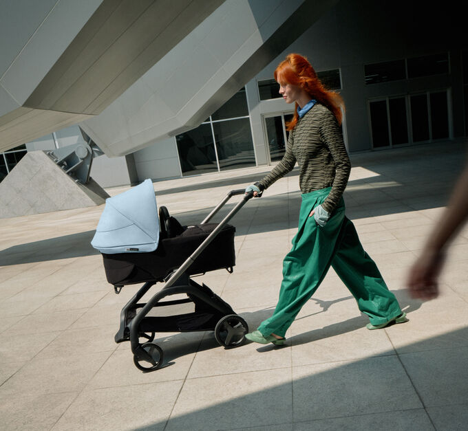 Una mamá segura de sí misma pasea con su bebé en un carrito urbano Bugaboo Dragonfly mientras pasa junto a un edificio futurista.