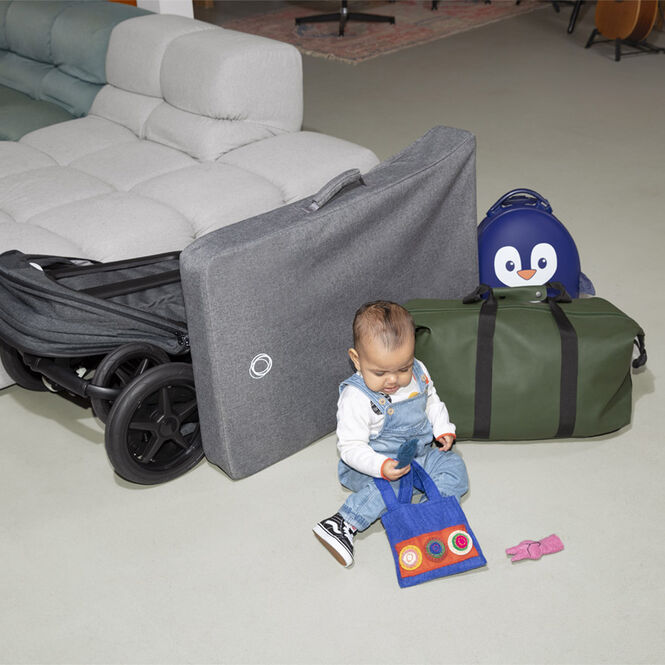 Baby playing on the floor, next to the folded Bugaboo Stardust in a carry bag and other travel luggage.