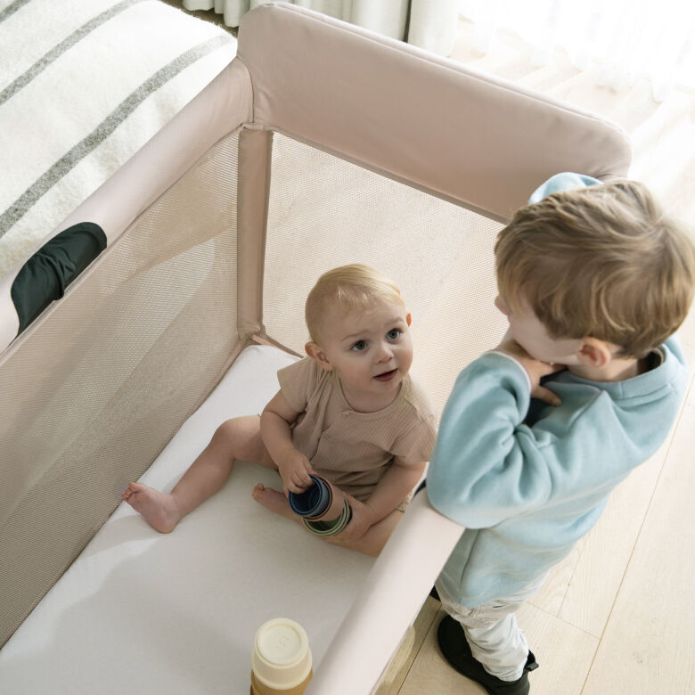 A baby sits in the Bugaboo Stardust and looks up at their brother, who is leaning over the frame of the travel cot.