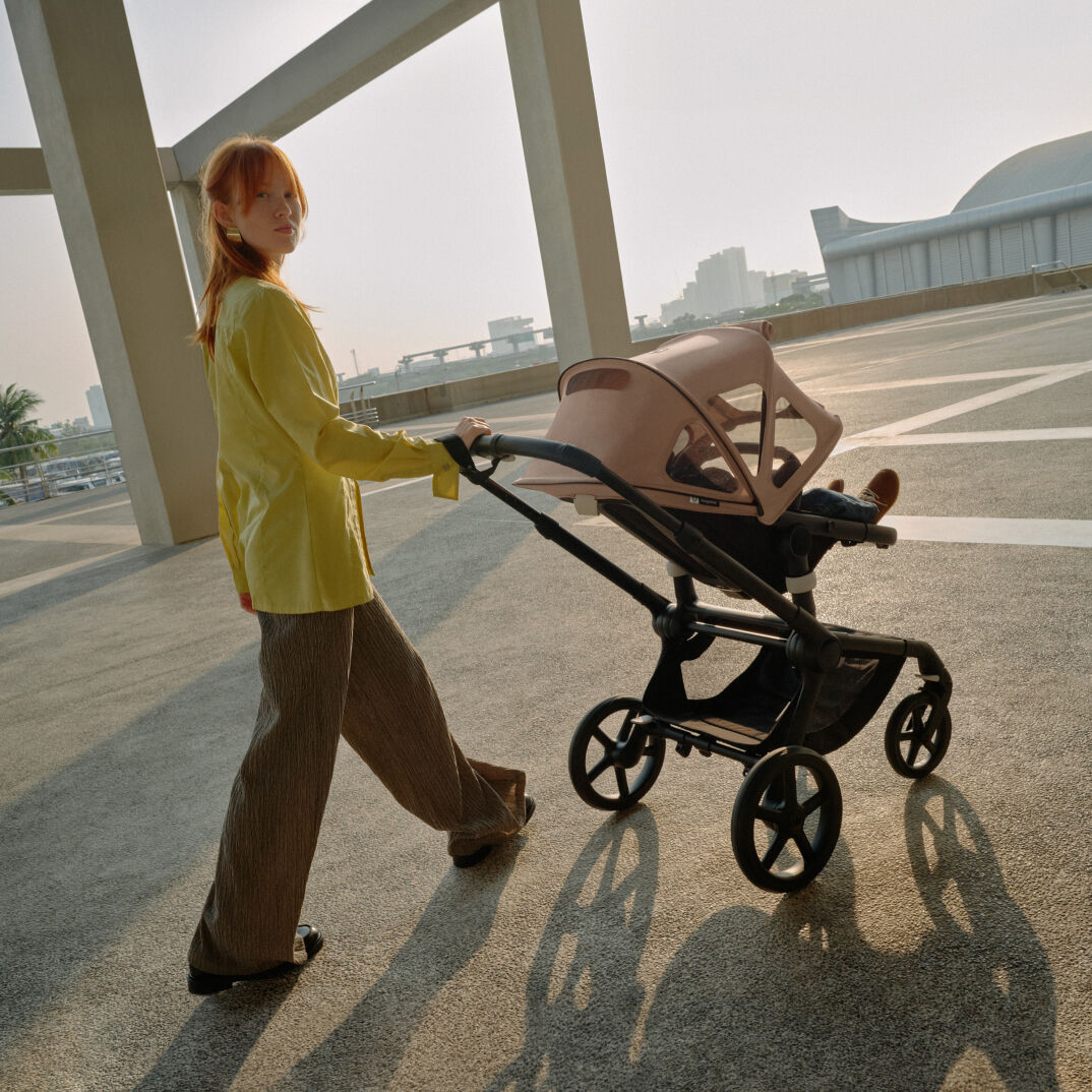 Een moeder kijkt naar de camera terwijl ze met haar baby in een Bugaboo Fox 5 wandelt. De wandelwagen heeft een roze breezy zonnekap.