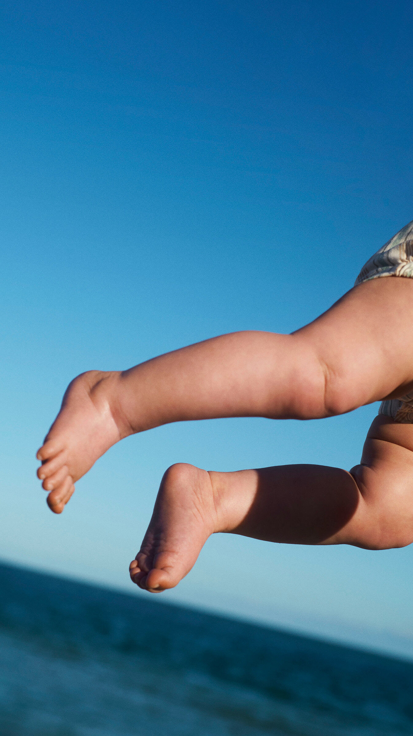 A close-up of a baby's legs and feet as she's thrown into the air, with the ocean and a clear blue sky in the background.