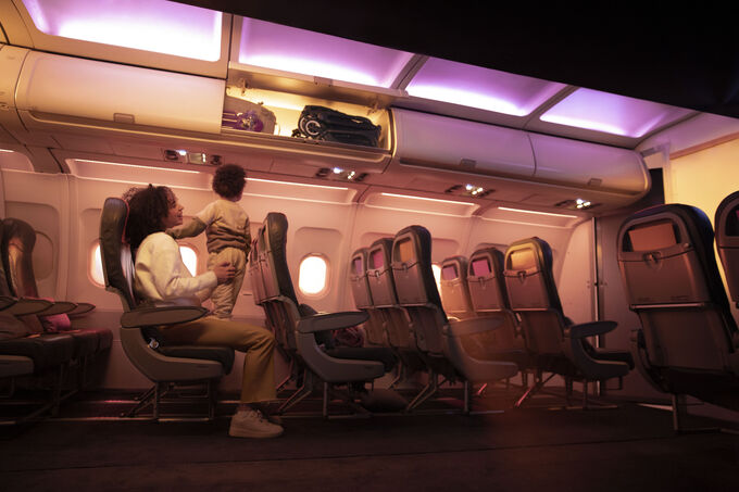Una mamá se sienta en el asiento de un avión con un niño de pie en su regazo; el niño mira el compartimiento superior de equipaje abierto con una silla de paseo urbana Bugaboo Butterfly plegada dentro