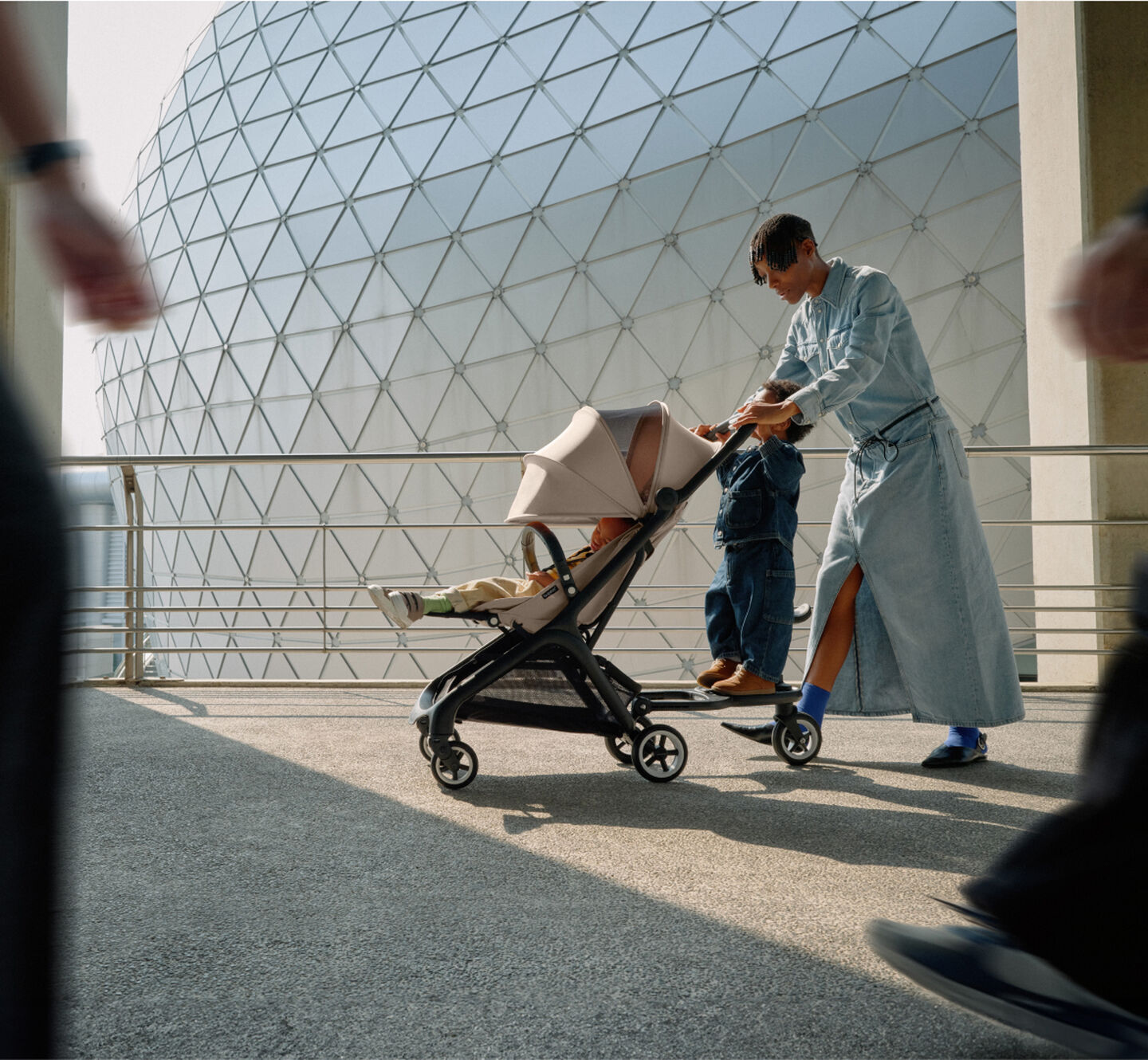 Eine stilvolle Mutter geht mit ihrem Baby in einem Bugaboo Butterfly-Reisekinderwagen spazieren, während ihr Kleinkind auf dem Rollbrett mitfährt.
