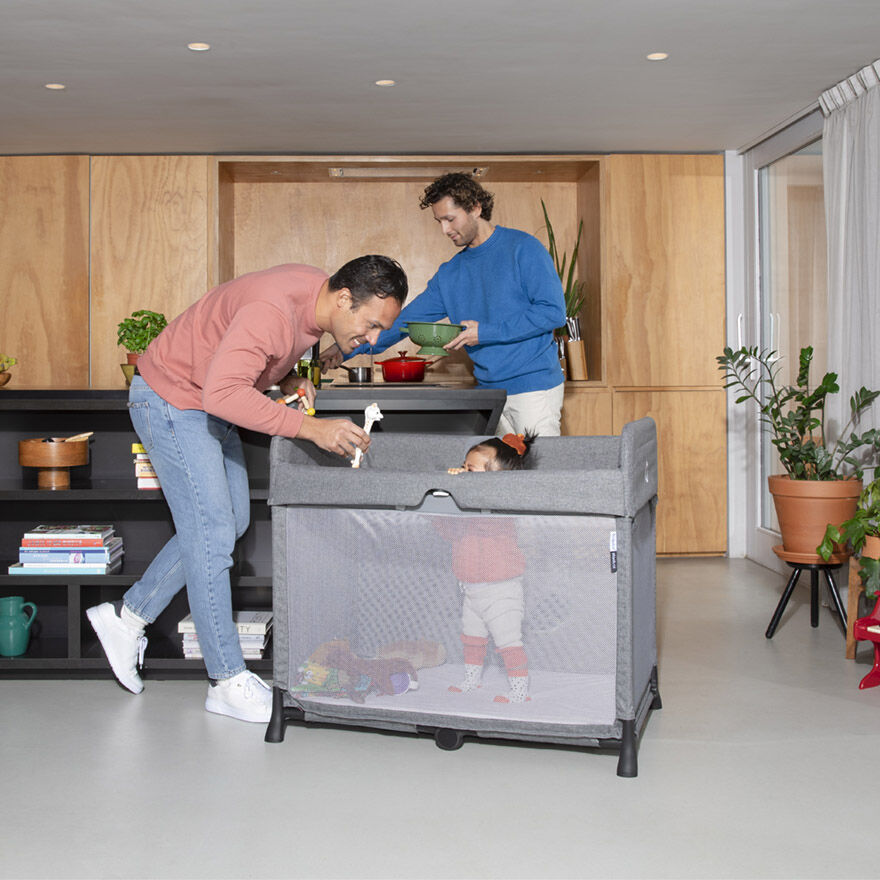 Two men are in the kitchen - one is cooking and the other is playing with a toddler, who is standing in the Bugaboo Stardust travel cot.