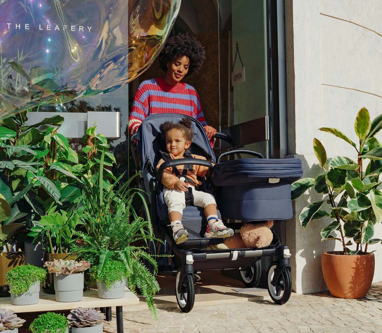 En mamma lämnar en butik och kör en Bugaboo Donkey 5 Duo med en sittdel och en liggdel. Ett barn sitter i sittdelen. Butikens entré är omgiven av grönska.
