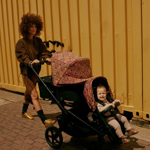 A stylish mom strolls confidently with a Bugaboo Kangaroo tandem stroller featuring a bassinet and a seat. One of her children sits facing forward in the seat. They’re walking alongside a wall of metal containers.