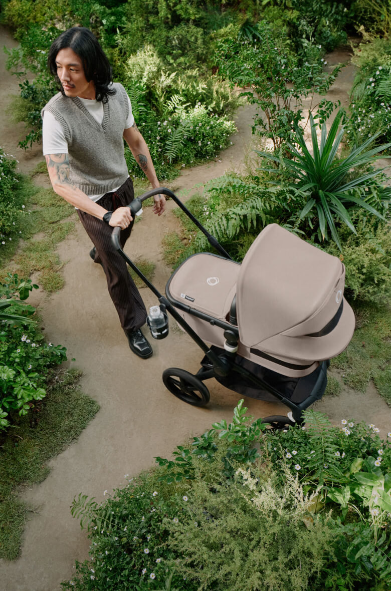 A dad takes a Bugaboo bassinet stroller through winding paths in a park, surrounded by lush greenery. The stroller features Desert Taupe fabrics and canopy.