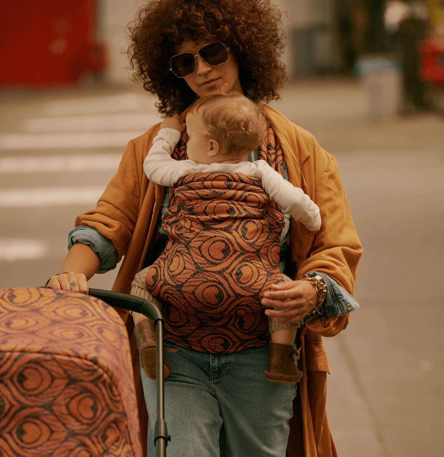 A stylish mom carries her baby in an Artipoppe Zeitgeist Baby Carrier with fabric that matches the Bugaboo stroller. Behind them is a casual, urban setting.