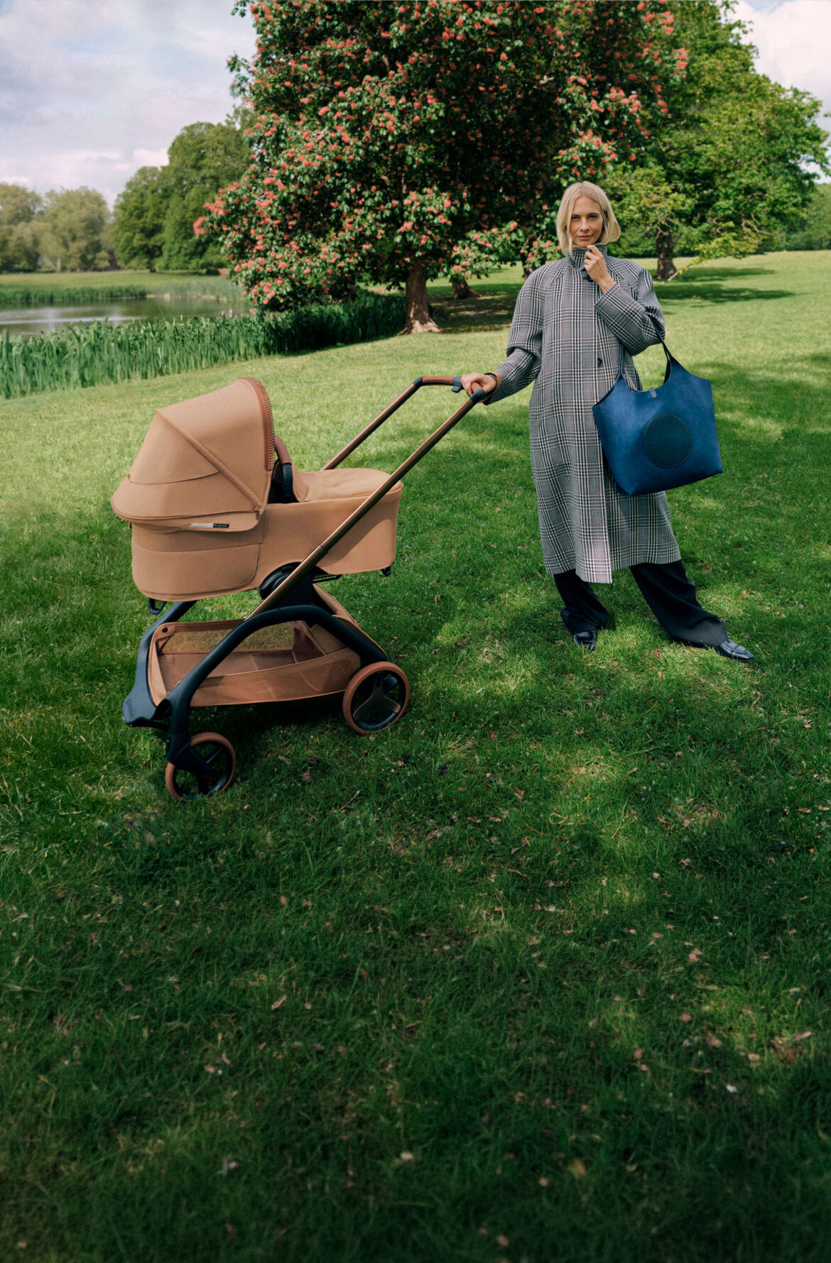 Poppy Delevingne stands next to a Bugaboo x Stella McCartney stroller, holding a large blue tote bag and wearing a long plaid coat. Behind her are a flowering tree, grassy fields, and a pond.