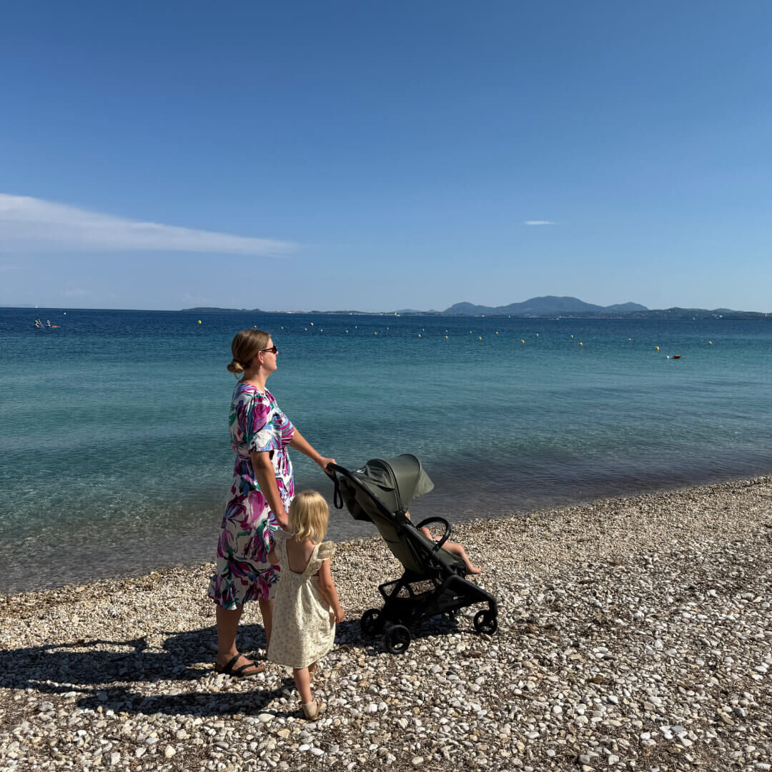 A mom walks hand in hand with her daughter on a pebbled beach while pushing a Bugaboo Butterfly 2 stroller with another child inside. The sea is calm and blue under a clear sky.