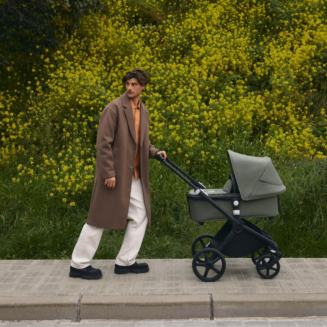 A stylish dad strolls along a walkway with a Bugaboo Fox Cub bassinet stroller in green fabrics. The slope behind him is covered in lush greenery and yellow wildflowers.