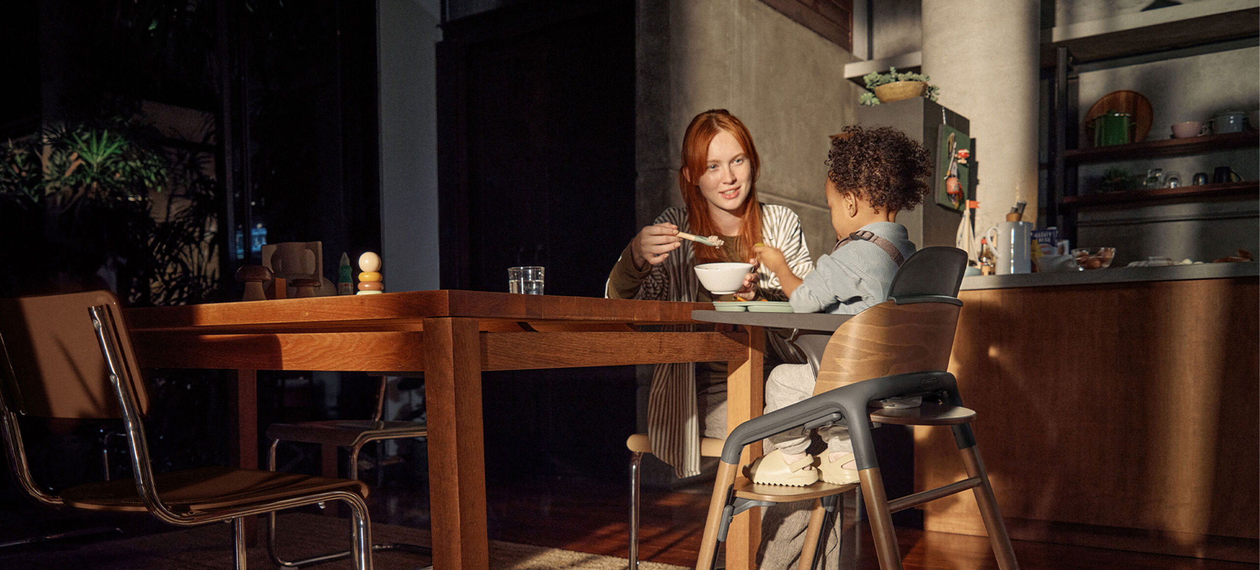 A mom feeds her toddler, who’s seated in a Bugaboo Giraffe high chair with a tray attached. They’re sitting in a warm, softly lit kitchen with wooden furniture.