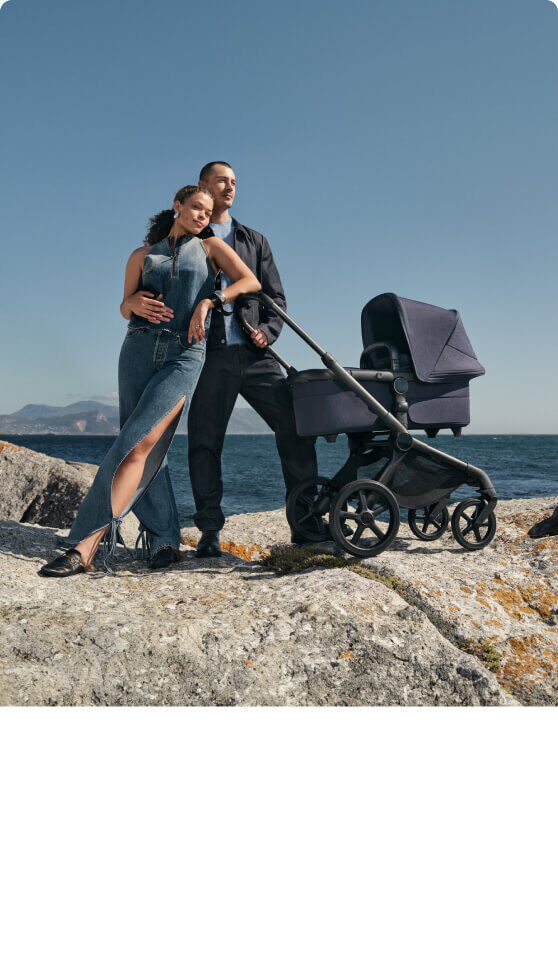 Two parents pose on a rocky coastline with the ocean in the background. Next to them is a sleek Bugaboo Fox 5 Renew newborn stroller in Heritage Black colorway.