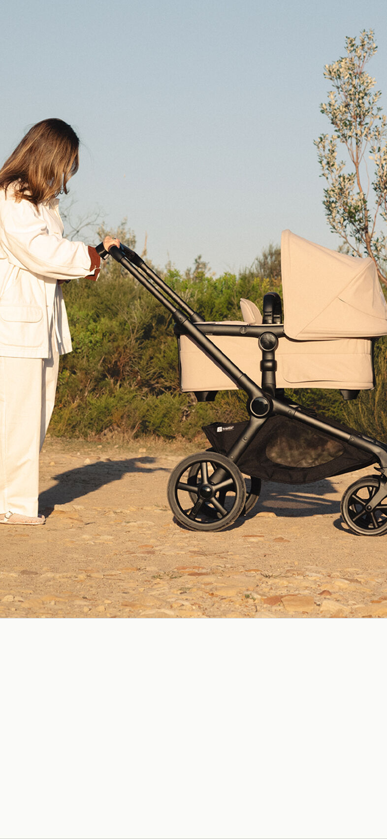 Parent pushing the Bugaboo Kangaroo pram in Desert Beige along a sunny countryside path surrounded by trees and greener.
