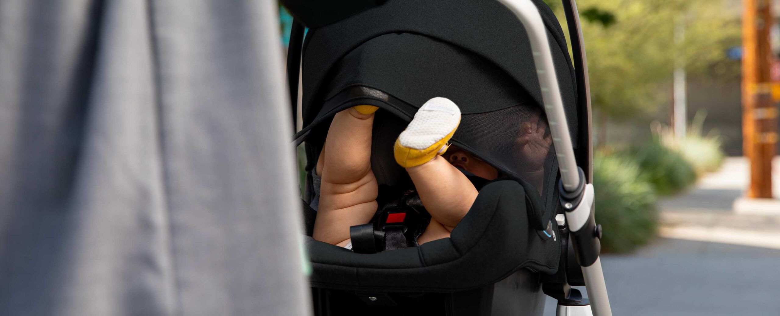 Un siège auto Bugaboo Turtle Air by Nuna fixé sur la poussette. Les pieds du bébé sont visibles, mais son corps est protégé par la capote. Un parent se tient au premier plan, flou, et l'arrière-plan montre un environnement extérieur composé d'un trottoir.