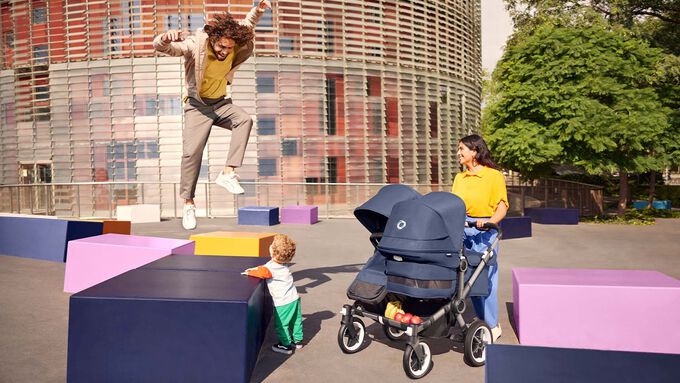 A family of four enjoying a day out; the father is carrying a Bugaboo changing backpack and pushing a Bugaboo pushchair, while a toddler is standing on a Bugaboo wheeled board.