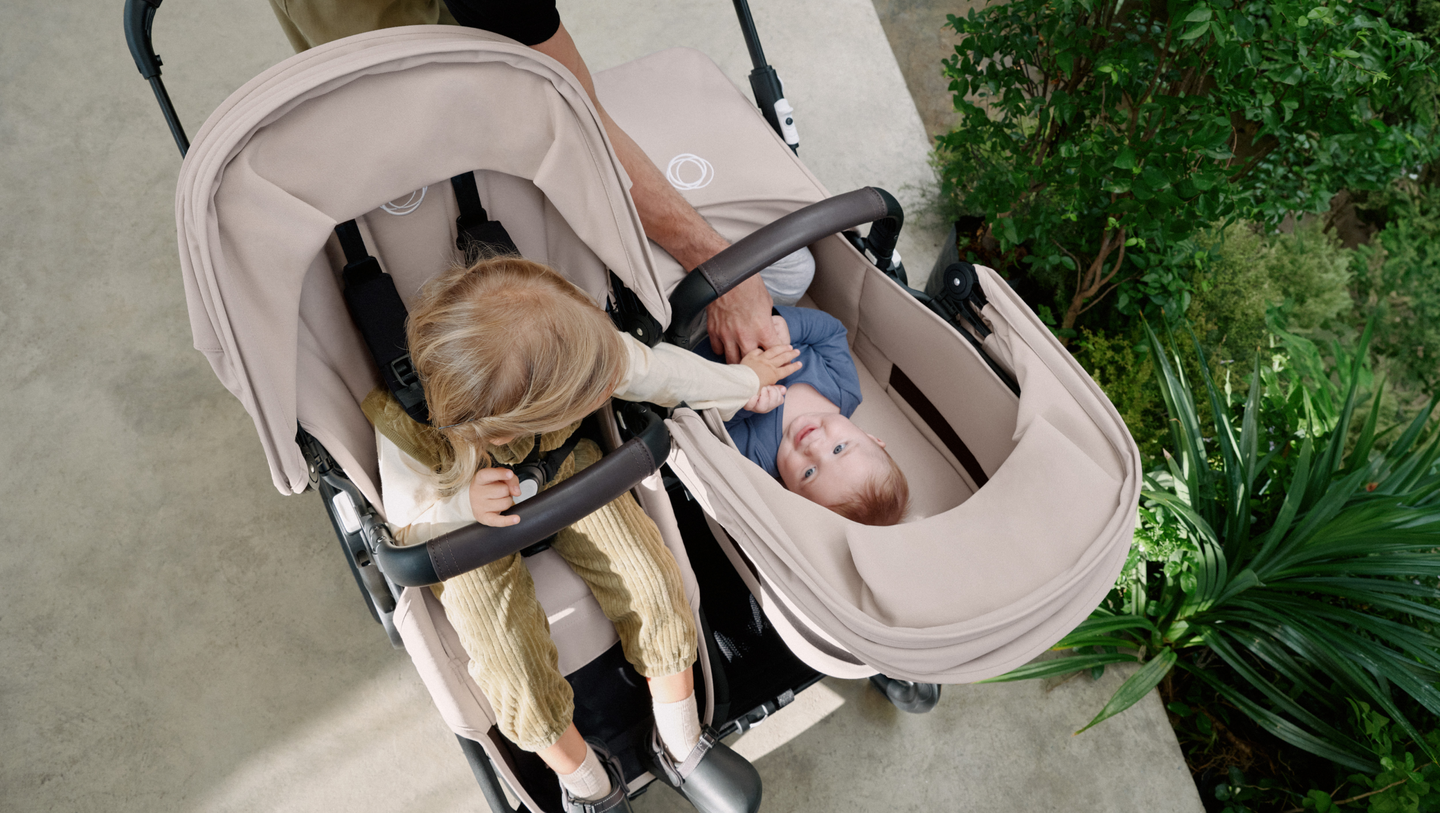 A mom strolls with two children in a Bugaboo Donkey 5 double stroller. The girl in the seat is reaching out to her sibling in the bassinet.