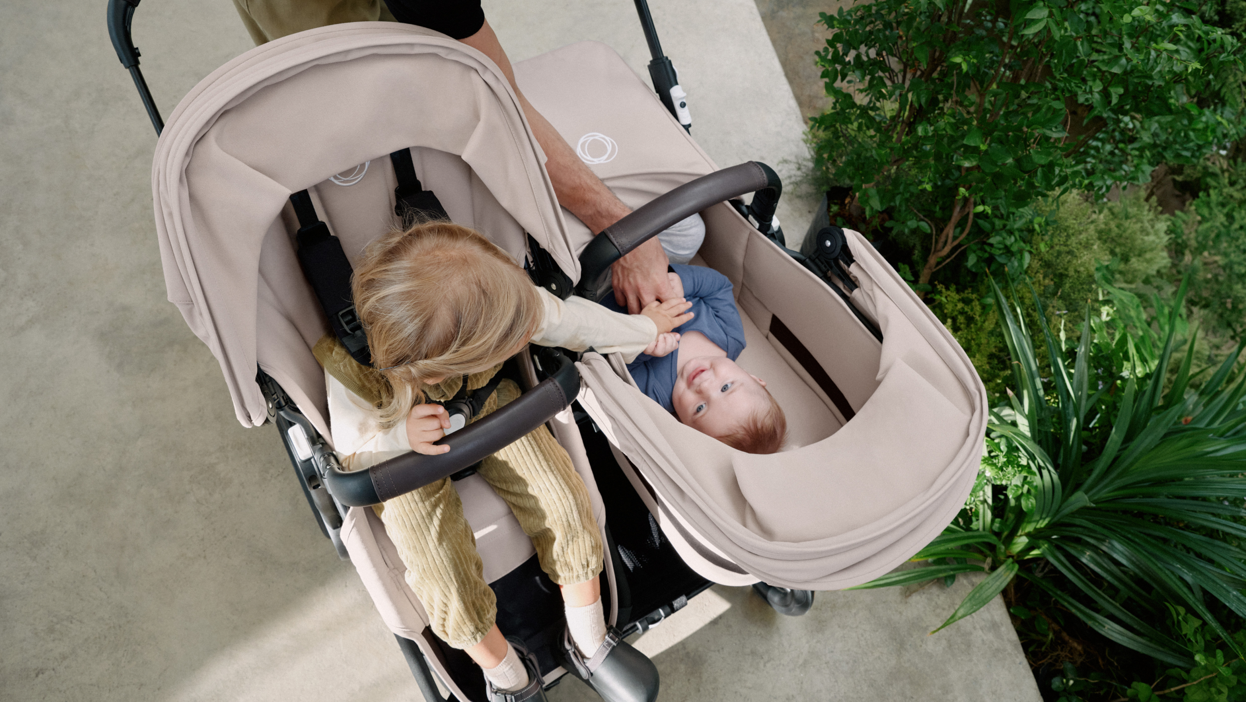 En mamma promenerar med två barn i en Bugaboo Donkey 5 syskonvagn. Flickan i sittdelen sträcker sig efter sitt syskon i liggdelen.