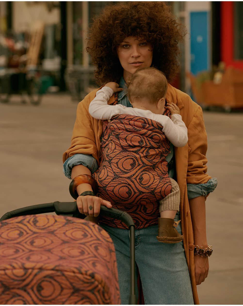 A stylish mom carries her baby in an Artipoppe Zeitgeist Baby Carrier with fabric that matches the Bugaboo stroller in front of them. The baby rests against her chest while she looks ahead calmly. Behind them is a casual, urban setting.