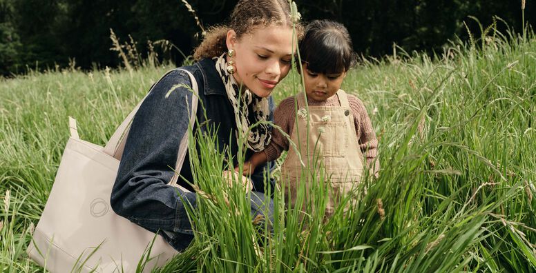 A mom and child crouch among tall grasses, curiously observing the plants. The mother carries a Bugaboo changing bag in Desert Taupe on her shoulder.