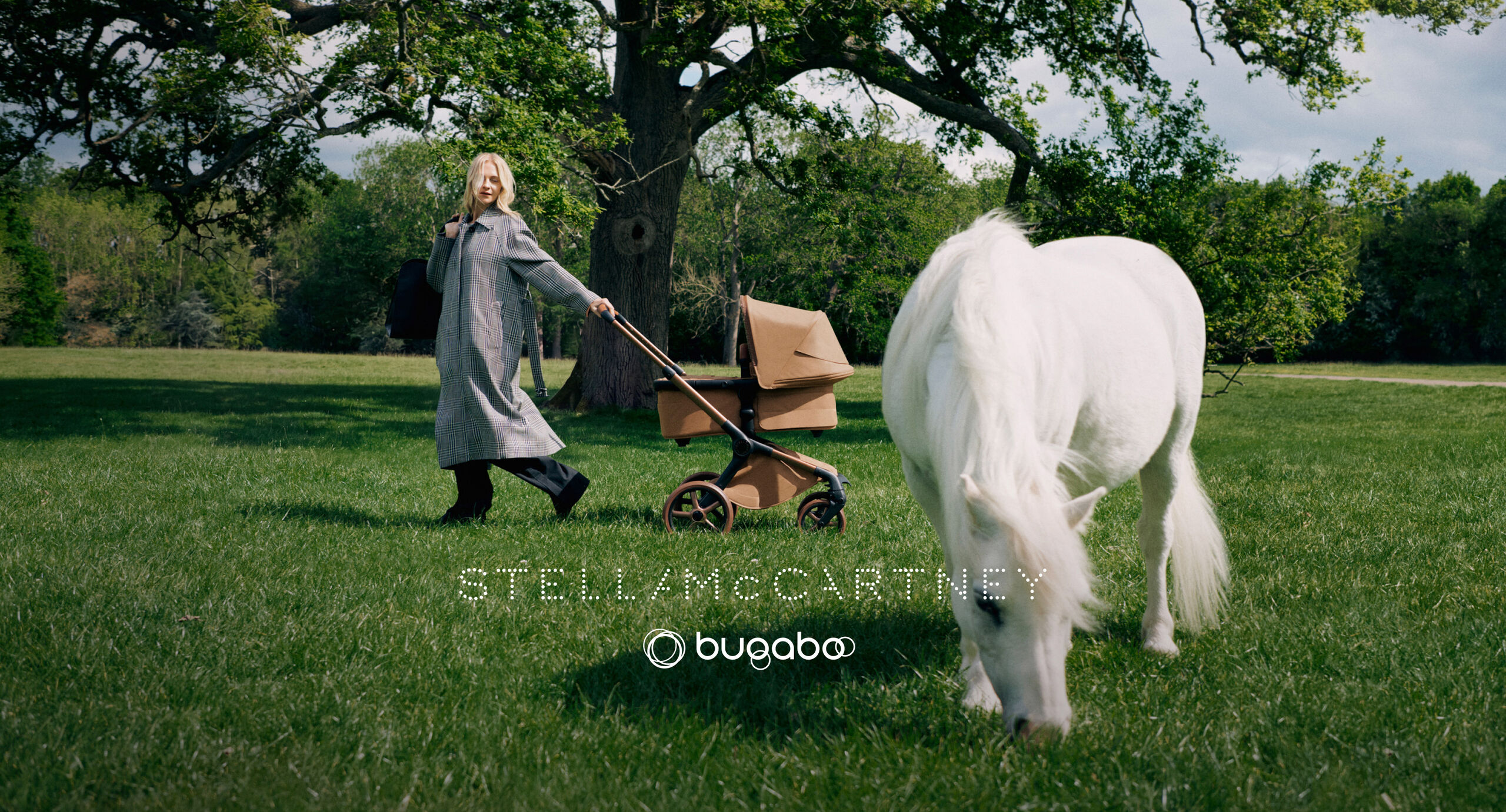 Poppy Delevingne se tient à côté d'une poussette Bugaboo x Stella McCartney, en tenant un grand sac fourre-tout bleu alors qu'elle porte un long manteau à carreaux. Un arbre en fleurs, des champs d'herbe et un étang se trouvent derrière elle.