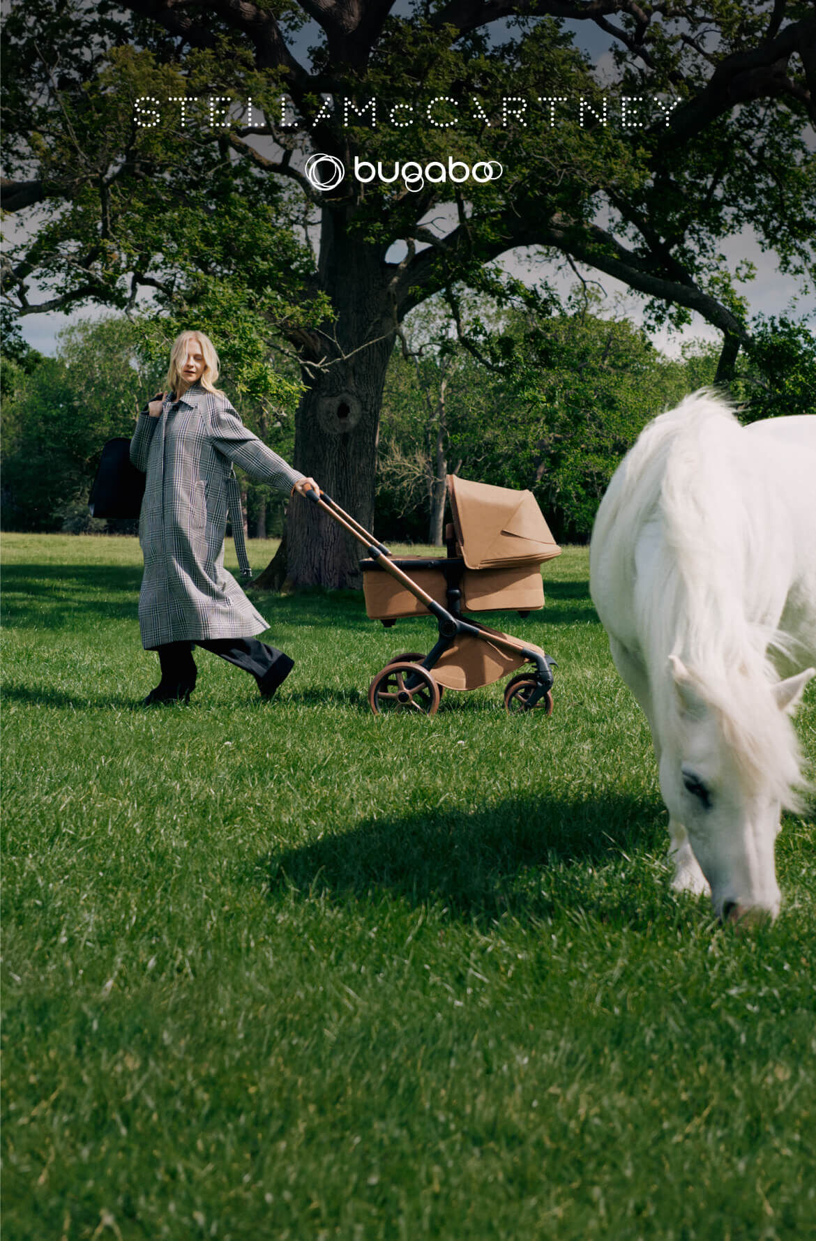 Poppy Delevingne se tient à côté d'une poussette Bugaboo x Stella McCartney, en tenant un grand sac fourre-tout bleu alors qu'elle porte un long manteau à carreaux. Un arbre en fleurs, des champs d'herbe et un étang se trouvent derrière elle.