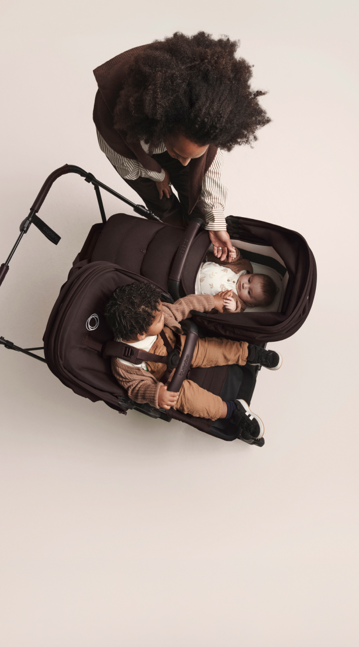 Top down view of the Bugaboo Donkey 6 stroller in double configuration. The toddler in the seat reaches out to his baby sibling in the bassinet. Their mom stands over them and gently touches the baby.