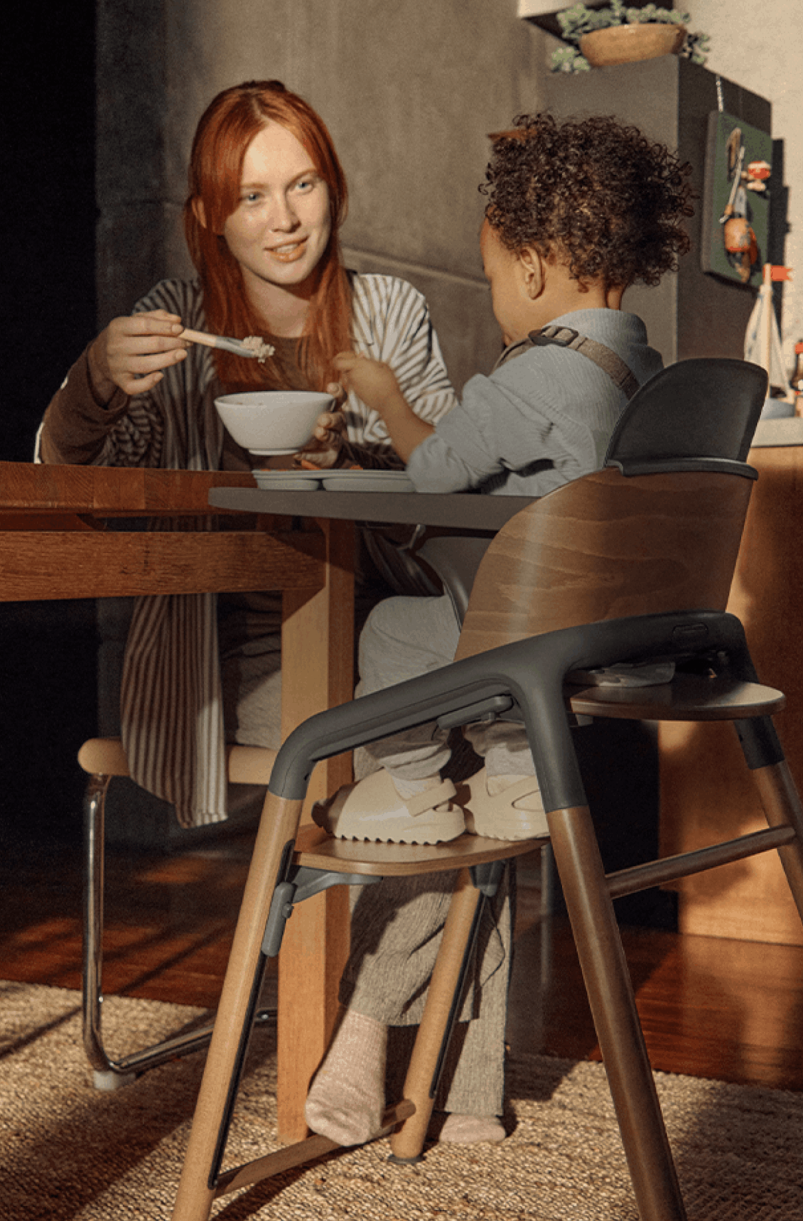 トレイが付いたバガブー ジラフ ハイチェアに座っている幼児に食事を与えている母親。 彼らは、木製家具に囲まれた、あたたかくやわらかな光が差し込むキッチンに座っている。