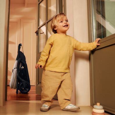 Compact folded Bugaboo stroller standing self-upright in a hallway next to a smiling toddler
