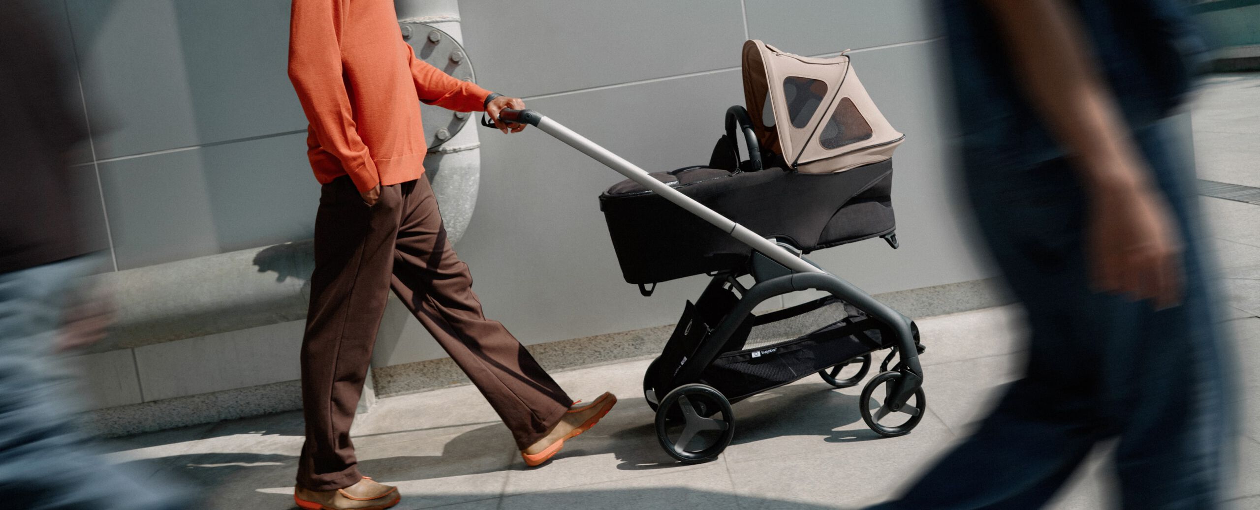 Un parent pousse une poussette Bugaboo avec une nacelle et une capote à fenêtres sur le trottoir. L'image est légèrement inclinée, avec des ombres floues en premier plan.