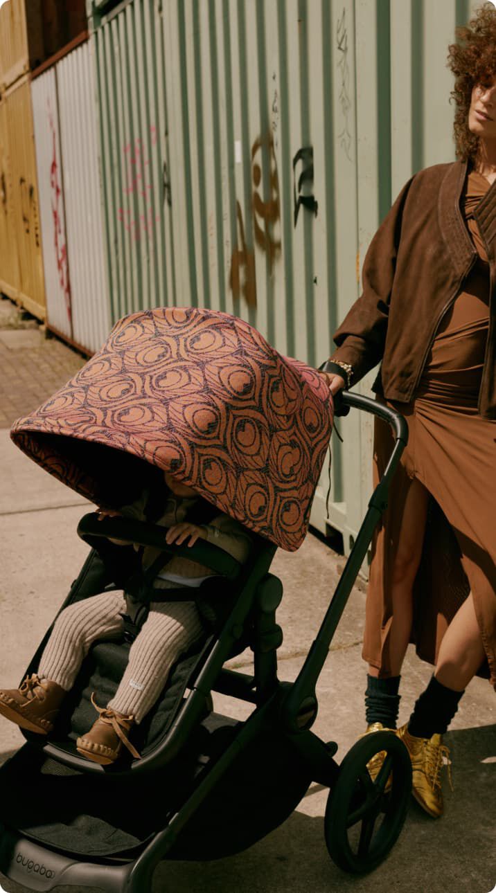 A stylish mom stands outdoors, leaning casually against the side of a metal container. Next to her, her child sits inside a Bugaboo Fox 5 Renew stroller with a Bugaboo X Artipoppe Limited Edition sun canopy.