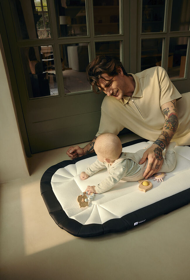 A dad gently supports his baby as she plays on a padded Bugaboo baby nest used as a playmat. They are at home, sitting together on the floor of a hallway.