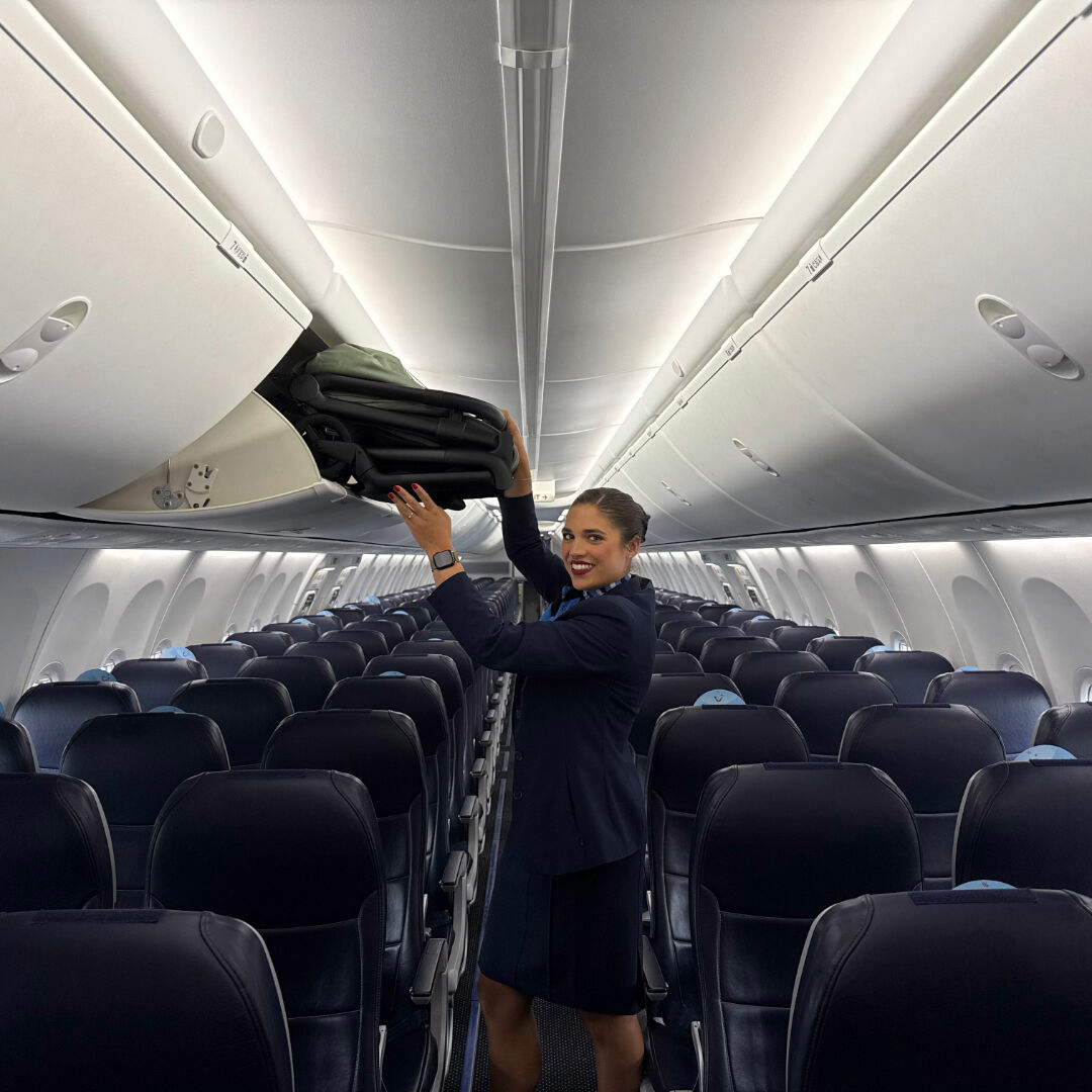 A smiling flight attendant stows a compactly folded Bugaboo Bugaboo 2 in the overhead compartment of an airplane.