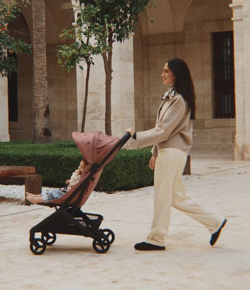 A smiling mom walks with her baby sitting in a Bugaboo Butterfly 2 stroller. The stroller features limited-edition fabric embroidered with Konges Sløjd cherry motif. They’re in a green courtyard outside a stone-walled building.