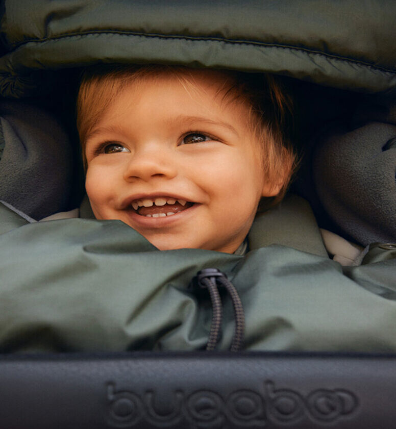 A baby smiling brightly as they're cuddled up in a footmuff inside a stroller.