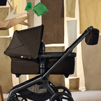 A Bugaboo Fox 5 Renew bassinet stroller in Cocoa Brown, with a Bugaboo organizer attached on the handlebar. The stroller stands in a stylized indoor set with a cardboard tree and abstract city-building shapes.