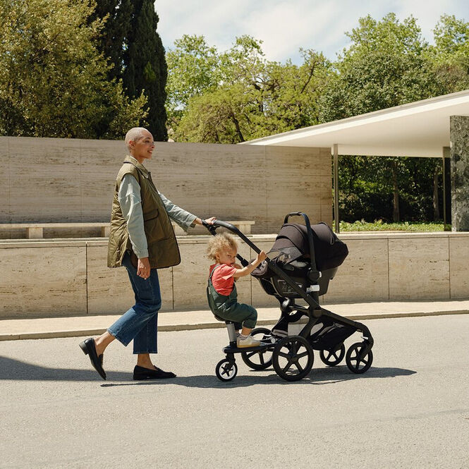 Een moeder wandelt met haar kinderen. Haar baby zit in de Bugaboo Fox 5 met een autostoel bevestigd. Haar oudere kind staat op het meerijdplankje.